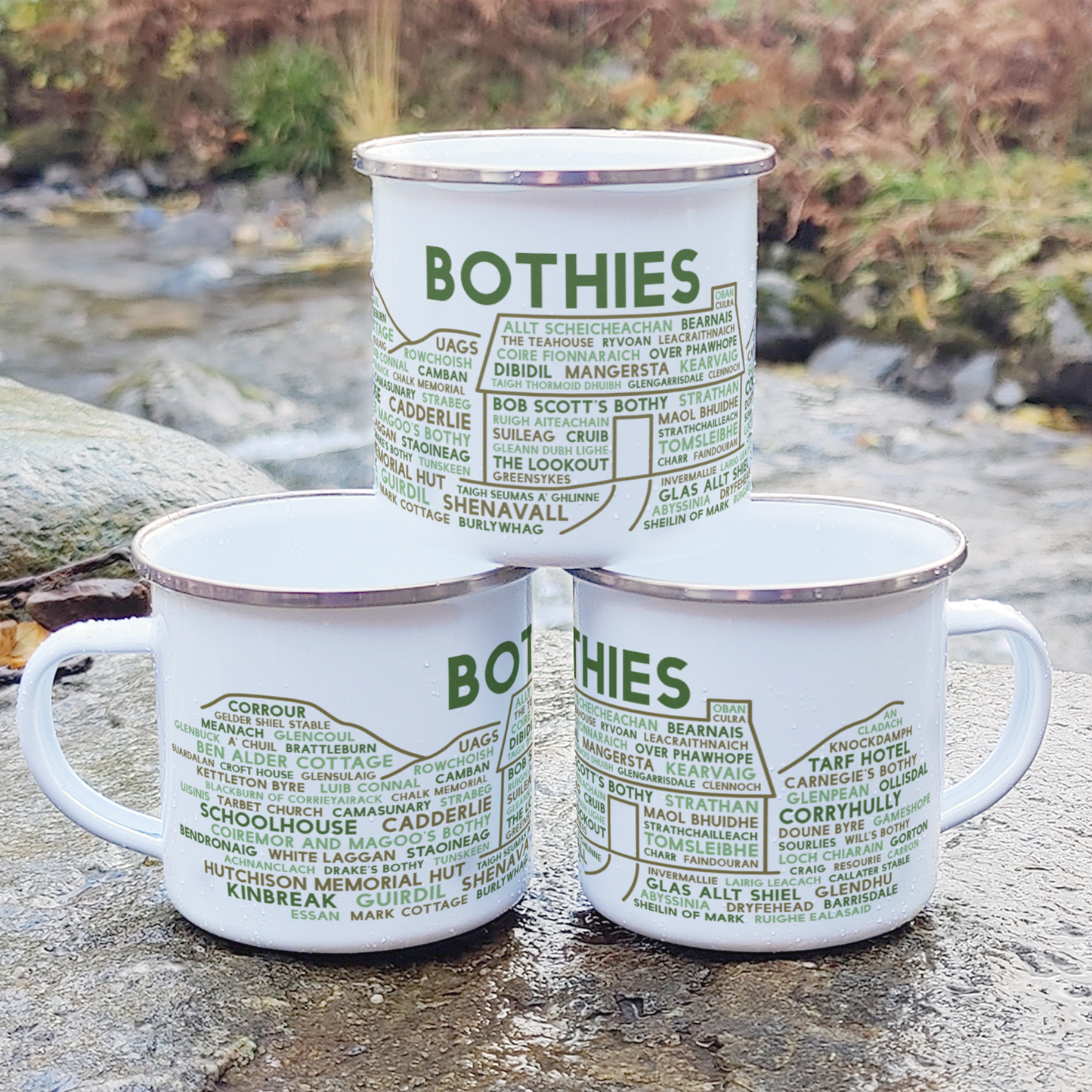 Three stacked white enamel mugs with silver rims, standing on a rock in front of foliage and a river. Each mug features the word "bothies" in green, with the names of various Scottish bothies underneath it.
