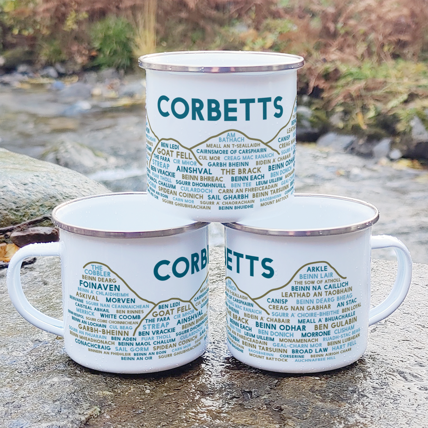 Three stacked white enamel mugs with silver rims, standing on a rock in front of foliage and a river. Each mug features the word "Corbetts" in blue, with the names of various Corbetts underneath it.