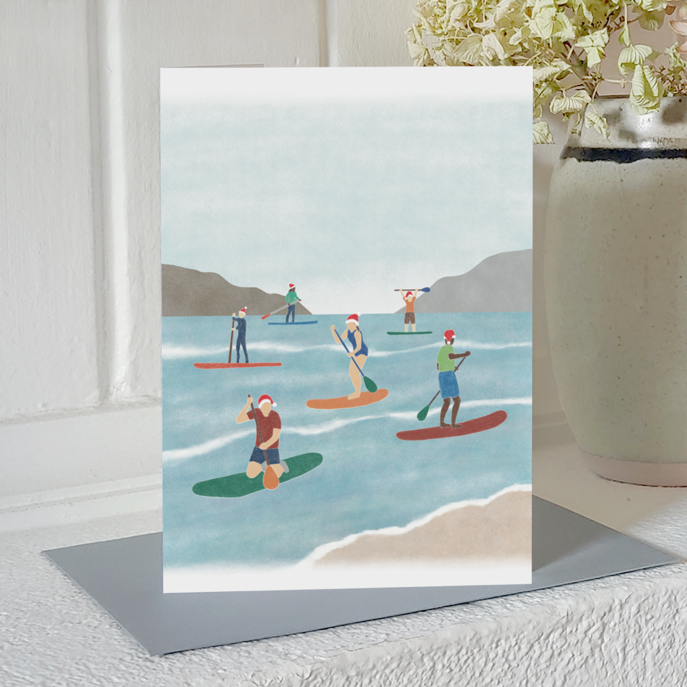 Paddle boarding Christmas card with an illustration of a group of paddle boarders wearing Santa hats while out on the water, standing on a mantelpiece