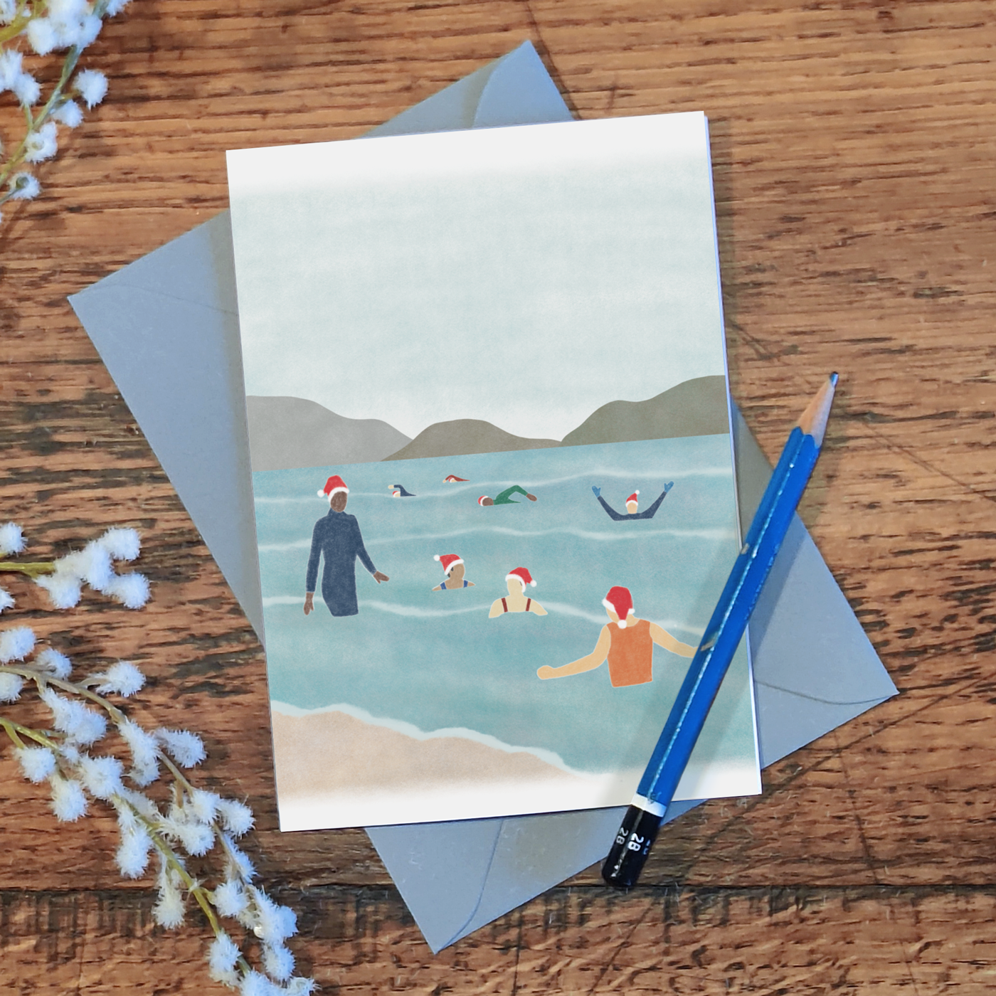 Wild swimming Christmas card with an illustration of a group of cold water swimmers wearing Santa hats, lying on a wooden surface with a pencil and some flowers