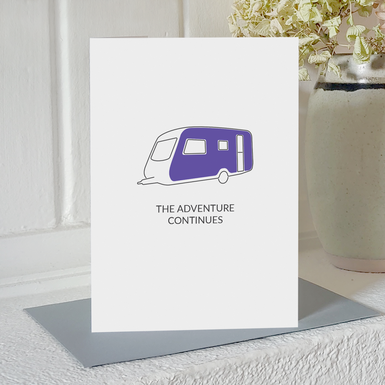 Card with illustration of a caravan and the message the adventure continues, standing on a white shelf