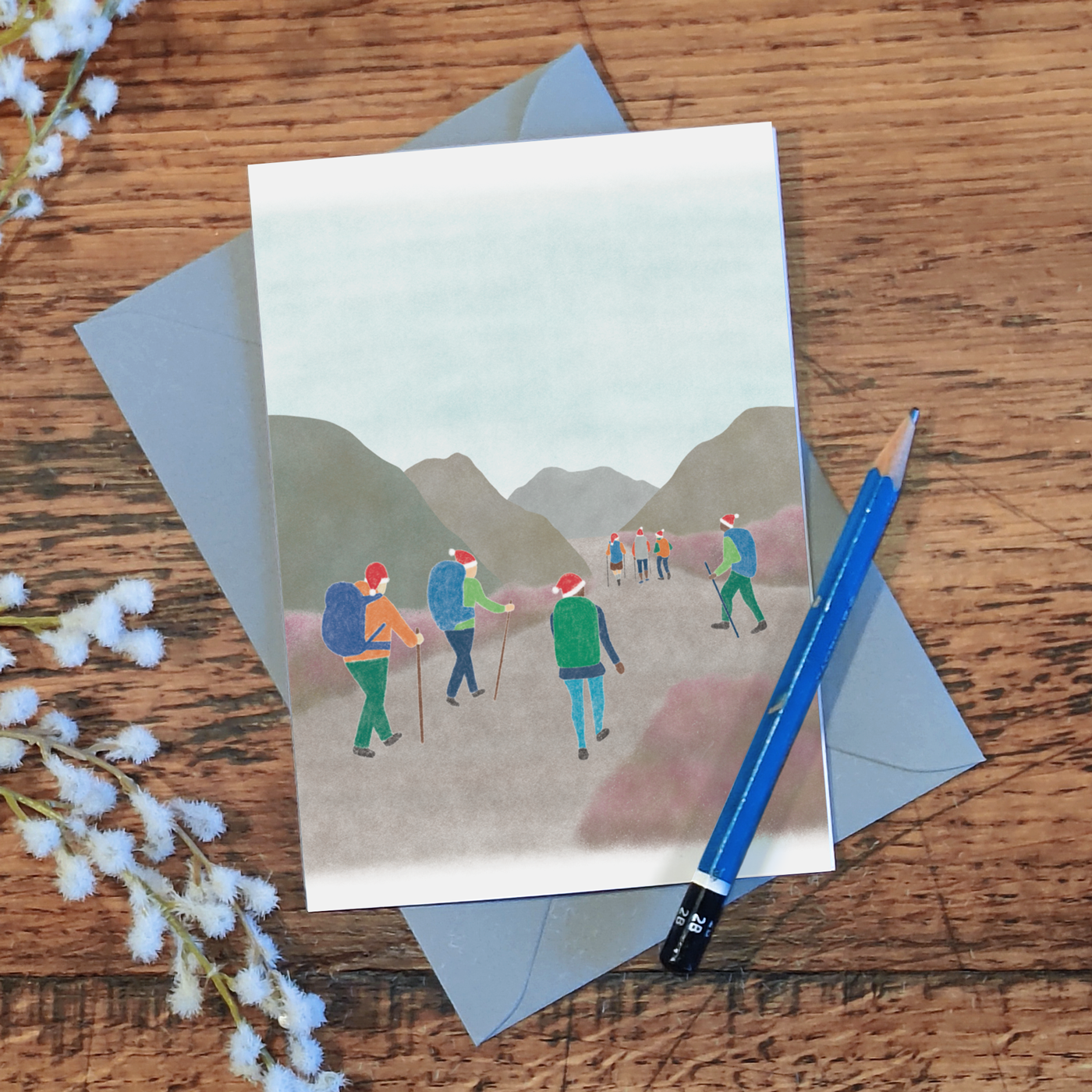 Hiking Christmas card with an illustration of a group of hikers wearing Santa hats, lying on a wooden surface with a pencil and some flowers
