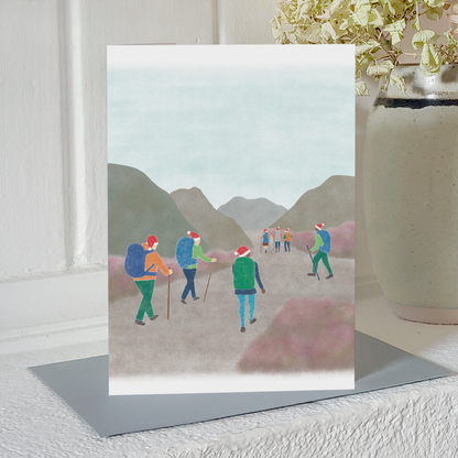 Hiking Christmas card with an illustration of a group of hikers wearing Santa hats, standing on a mantelpiece