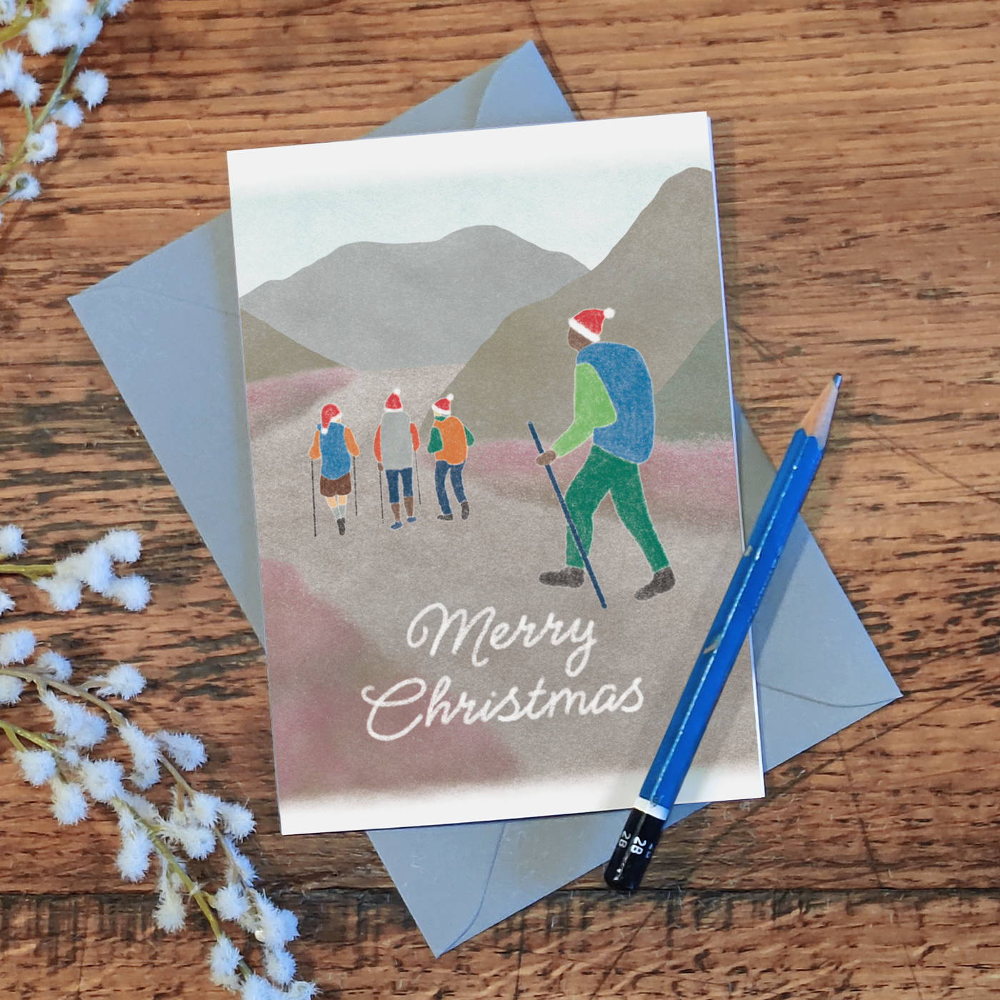 Christmas card with illustration of people wearing Santa hats hiking in the mountains and the text 'Merry Christmas', lying on a wooden surface with a pencil and flowers