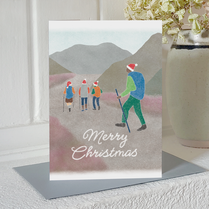 Christmas card with illustration of people wearing Santa hats hiking in the mountains and the text 'Merry Christmas', standing on a mantelpiece