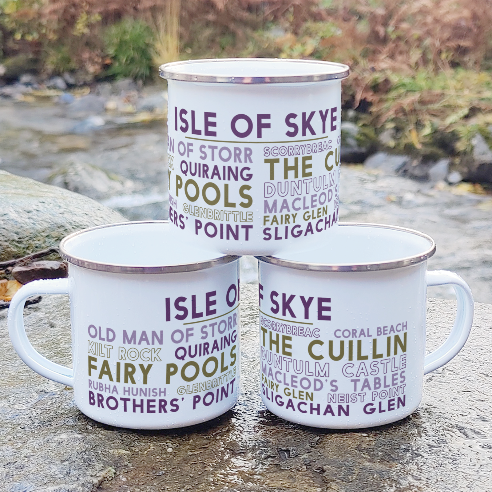 Three stacked white enamel mugs with silver rims, standing on a rock in front of foliage and a river. Each mug features the words "Isle of Skye" in purple, with the names of various landmarks and attractions on Skye underneath