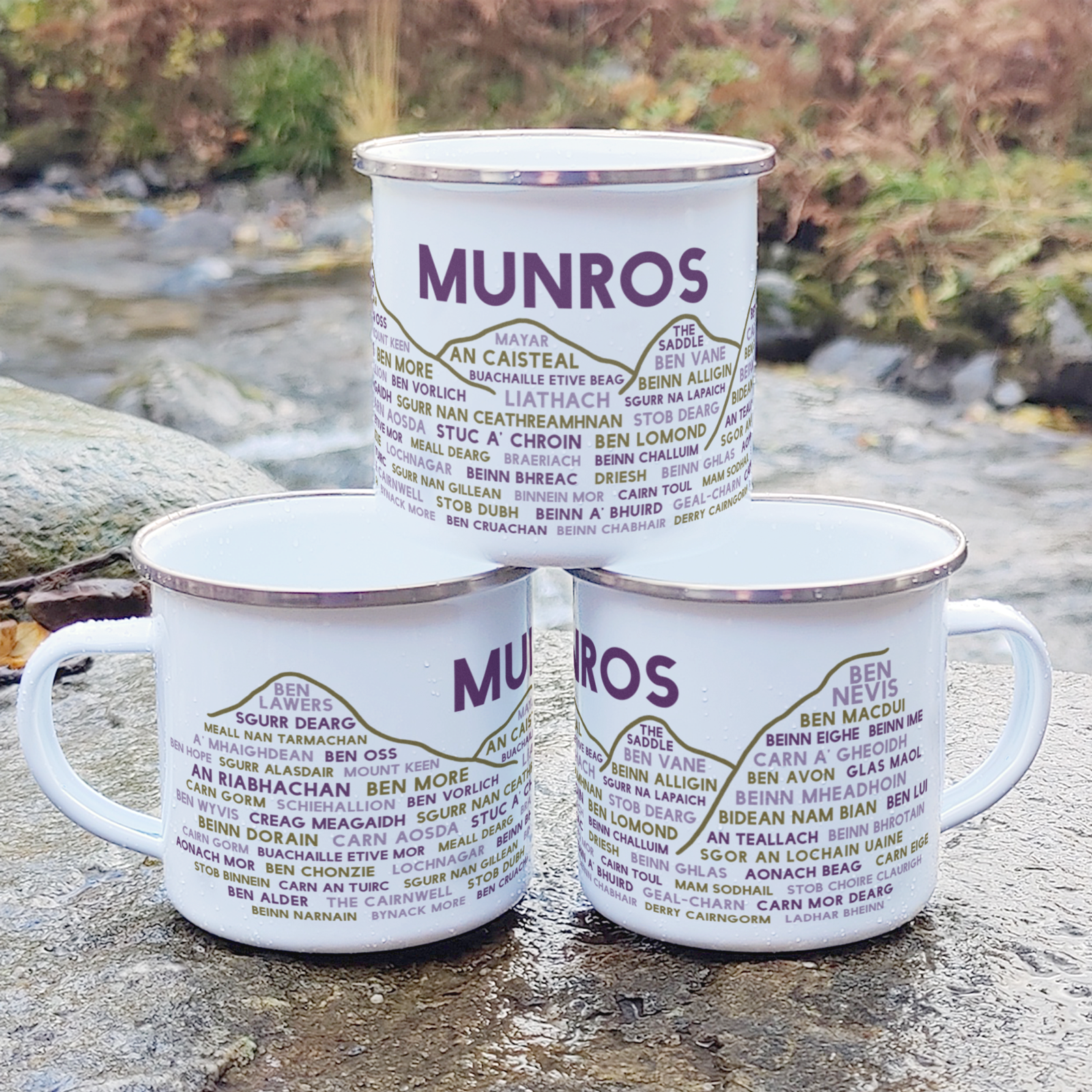 Three stacked white enamel mugs with silver rims, standing on a rock in front of foliage and a river. Each mug features the word "Munros" in purple, with the names of various Munros underneath it.