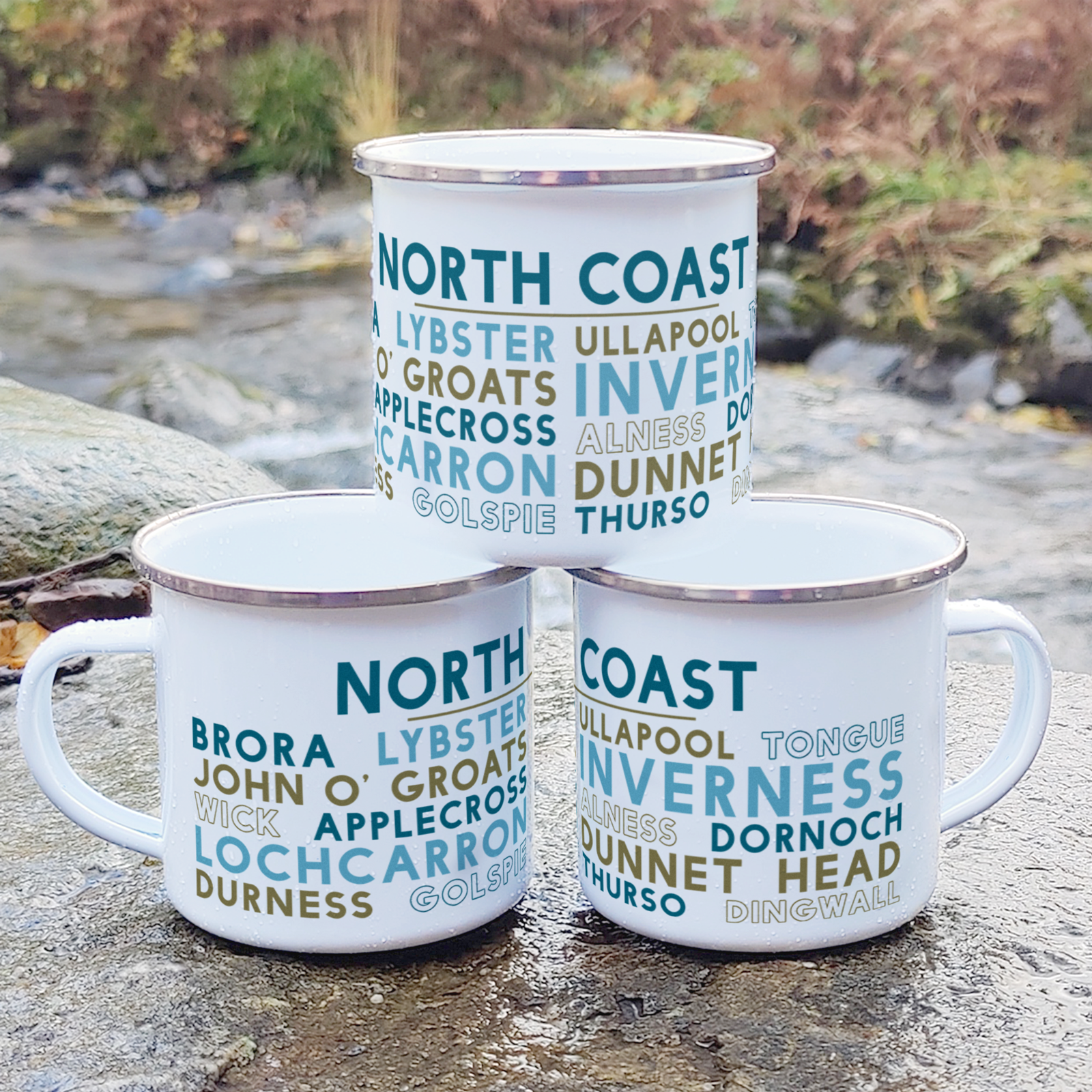 Three stacked white enamel mugs with silver rims, standing on a rock in front of foliage and a river. Each mug features the words "North Coast" in blue, with the names of various towns around the North Coast of Scotland underneath