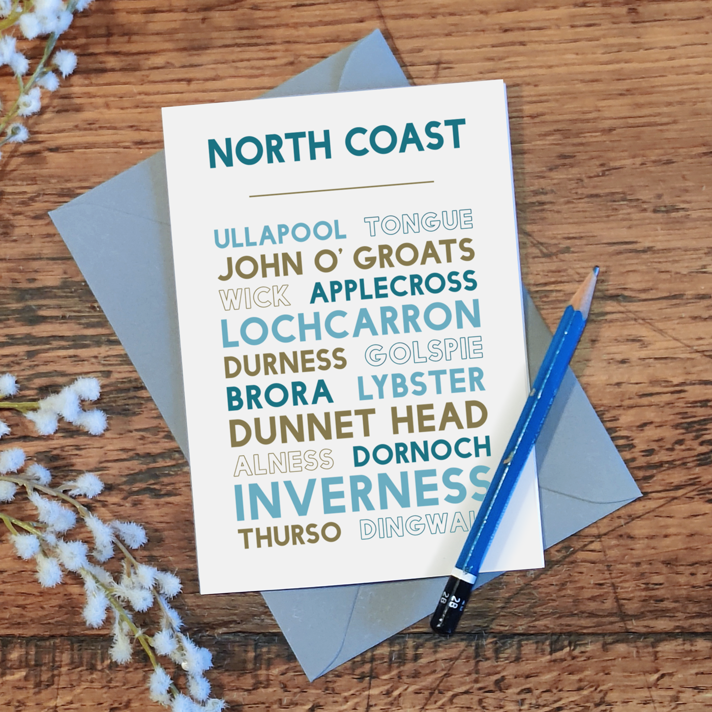 North Coast of Scotland greetings card lying on a wooden table with a pencil and flowers