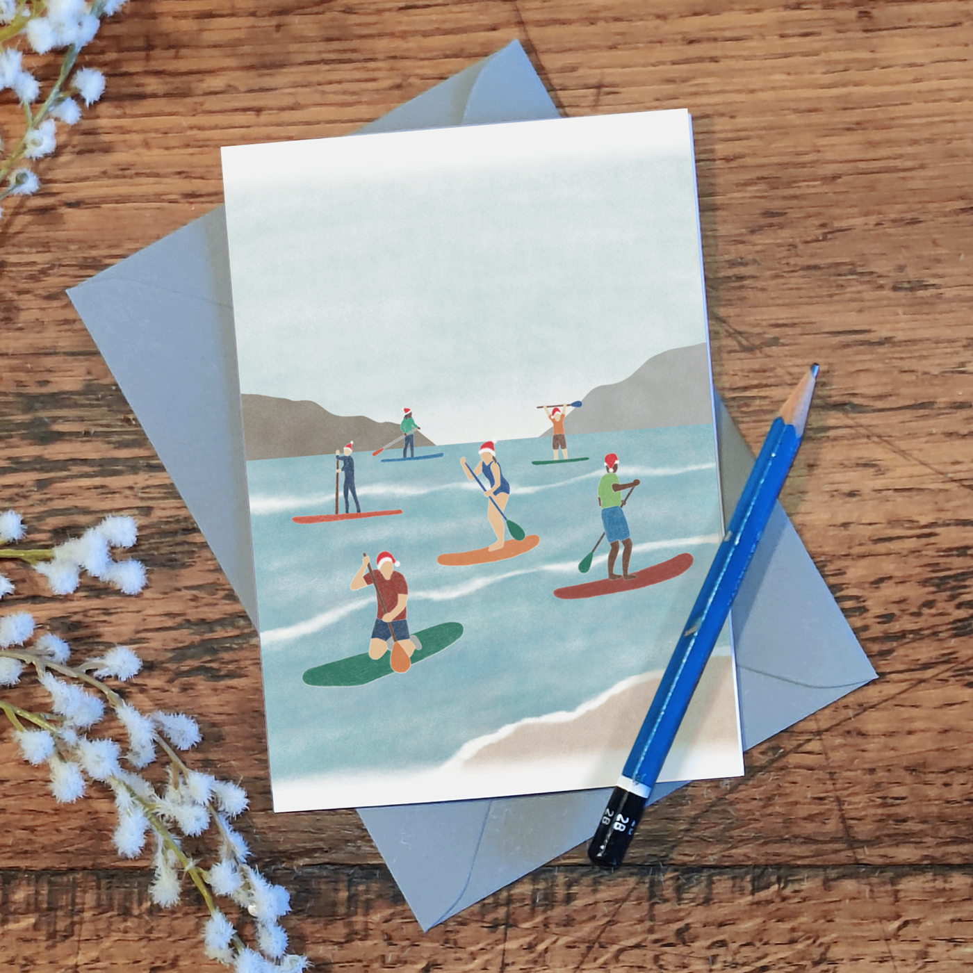 Paddle boarding Christmas card with an illustration of a group of paddle boarders wearing Santa hats while out on the water, on a wooden surface with a pencil and some flowers