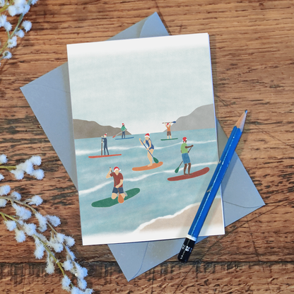 Paddle boarding Christmas card with an illustration of a group of paddle boarders wearing Santa hats while out on the water, on a wooden surface with a pencil and some flowers