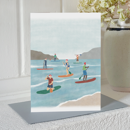 Paddle boarding Christmas card with an illustration of a group of paddle boarders wearing Santa hats while out on the water, standing on a mantelpiece