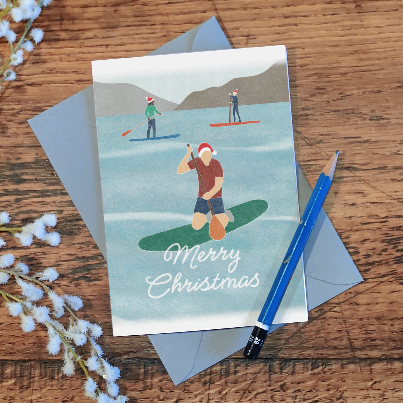 Paddle boarding Christmas card with three paddleboarders wearing Santa hats and the message "Merry Christmas", lying on a wooden surface with a pencil and some flowers