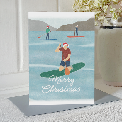 Paddle boarding Christmas card with three paddleboarders wearing Santa hats and the message "Merry Christmas", standing on a mantelpiece