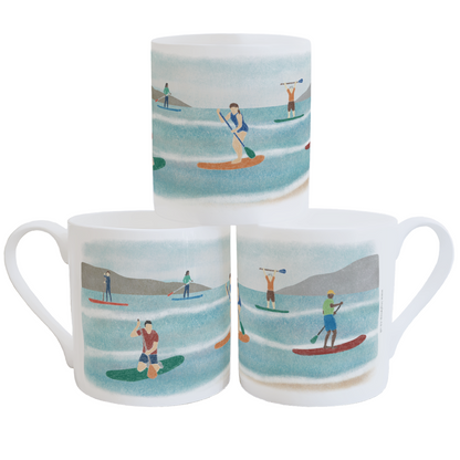 A stack of three white china mugs, decorated with a colourful illustration of people using paddleboards