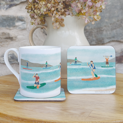 A fine bone china mug sitting on a coaster, with a second coaster stood next to it, all featuring an illustration of paddleboarders