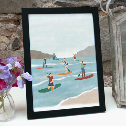 Paddleboarding print - an illustration of six paddle boarders out on the water - in a black frame standing on a shelf