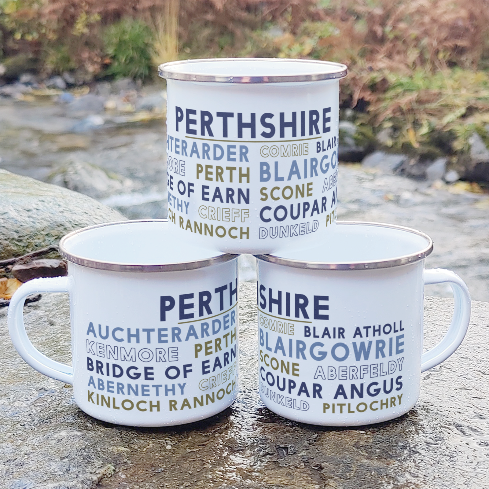 Three stacked white enamel mugs with silver rims, standing on a rock in front of foliage and a river. Each mug features the word "Perthshire" in purple, and the names of various towns and cities in Perthshire underneath