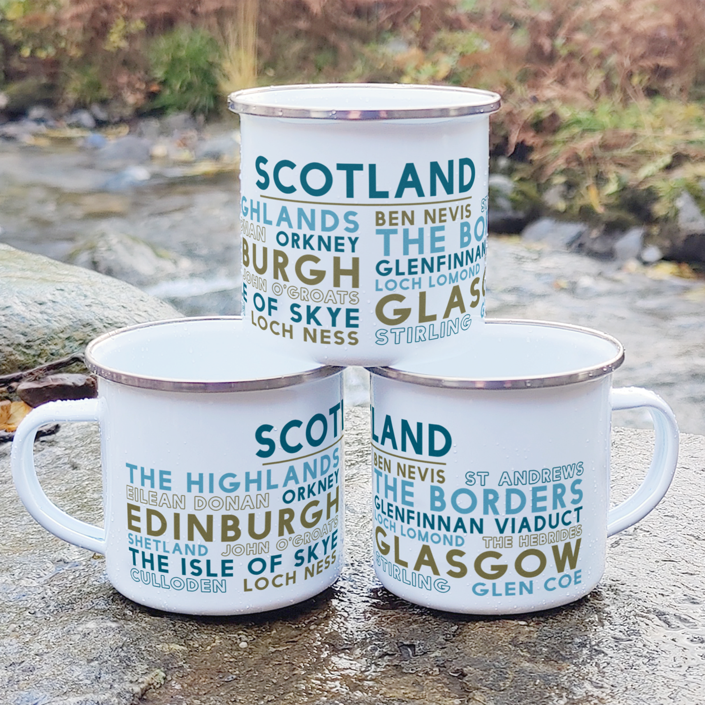 Three stacked white enamel mugs with silver rims, standing on a rock in front of foliage and a river. Each mug features the word "Scotland" in blue, with the names of famous towns and landmarks in Scotland underneath