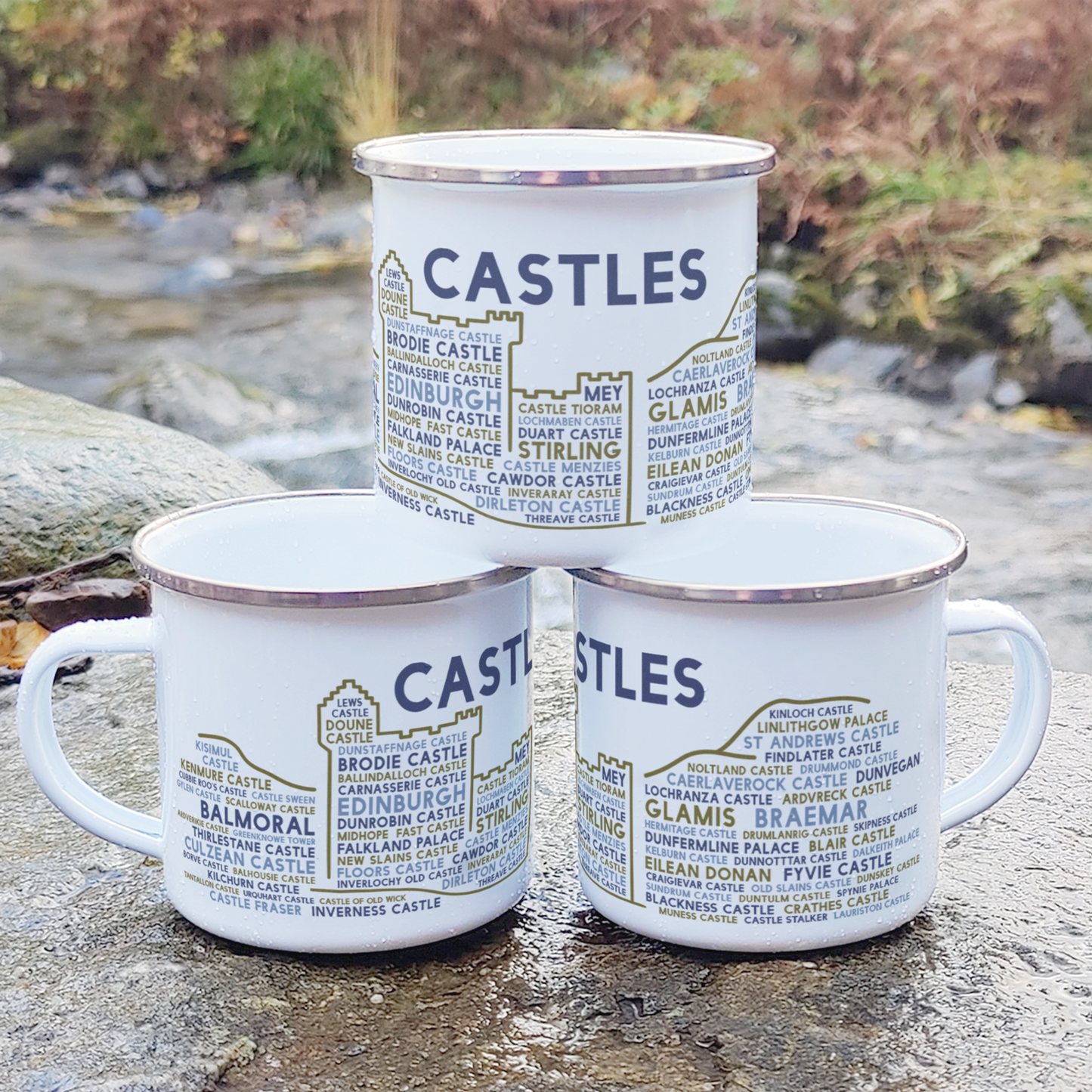 Three stacked white enamel mugs with silver rims, standing on a rock in front of foliage and a river. Each mug features the word "castles" in purple, with the names of various Scottish castles underneath it.