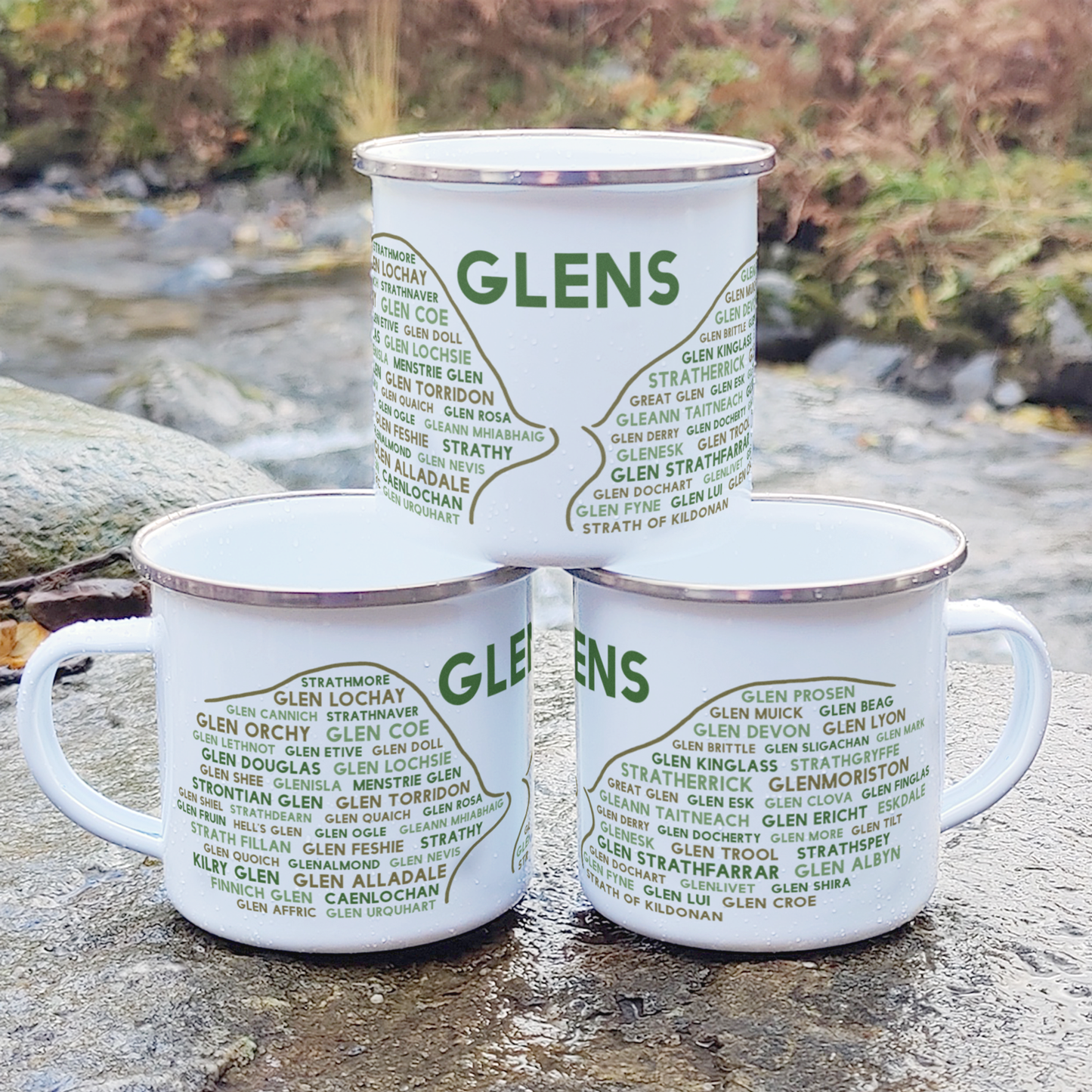 Three stacked white enamel mugs with silver rims, standing on a rock in front of foliage and a river. Each mug features the word "glens" in green, with the names of various Scottish glens underneath it.