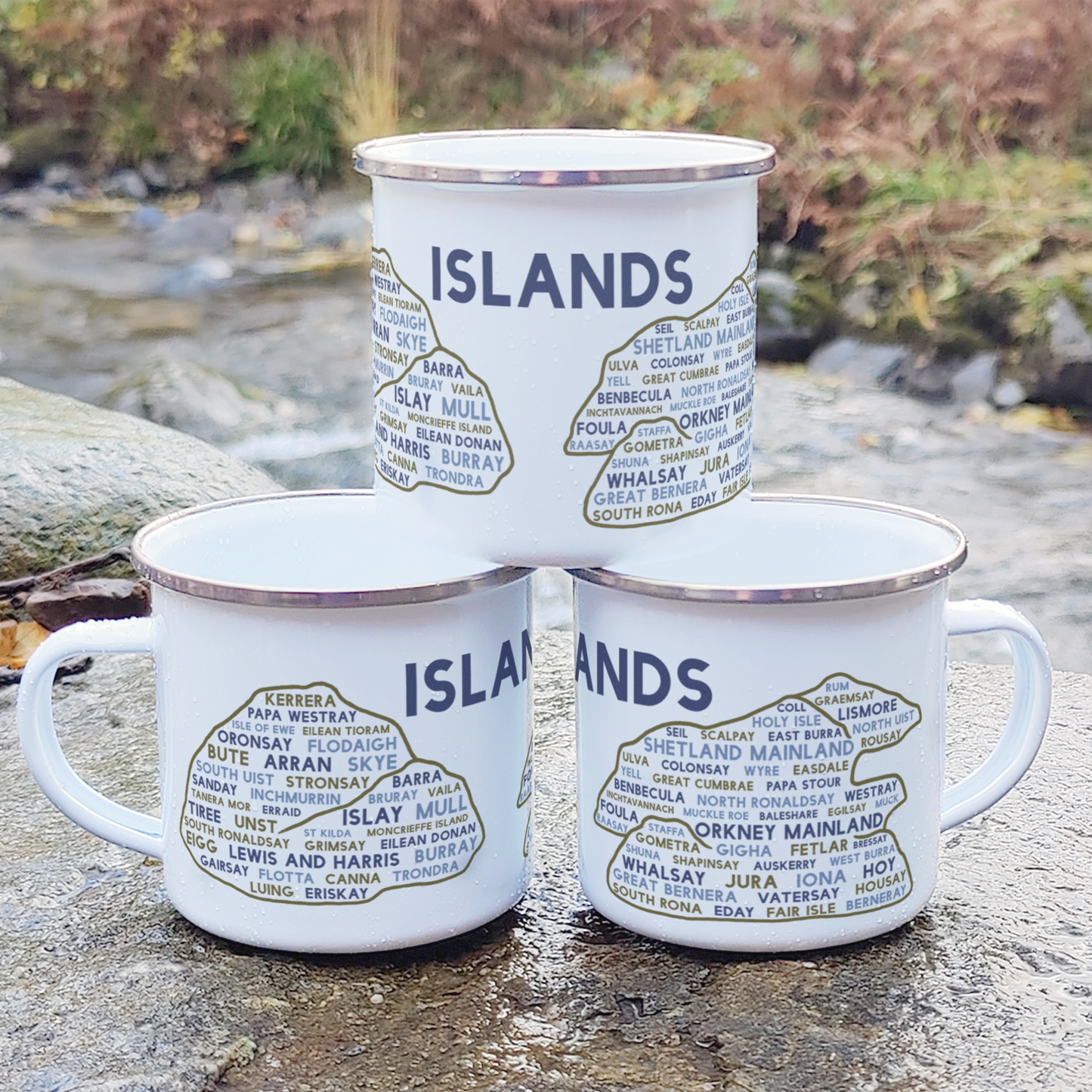 Three stacked white enamel mugs with silver rims, standing on a rock in front of foliage and a river. Each mug features the word "islands" in purple, with the names of various Scottish islands underneath it.