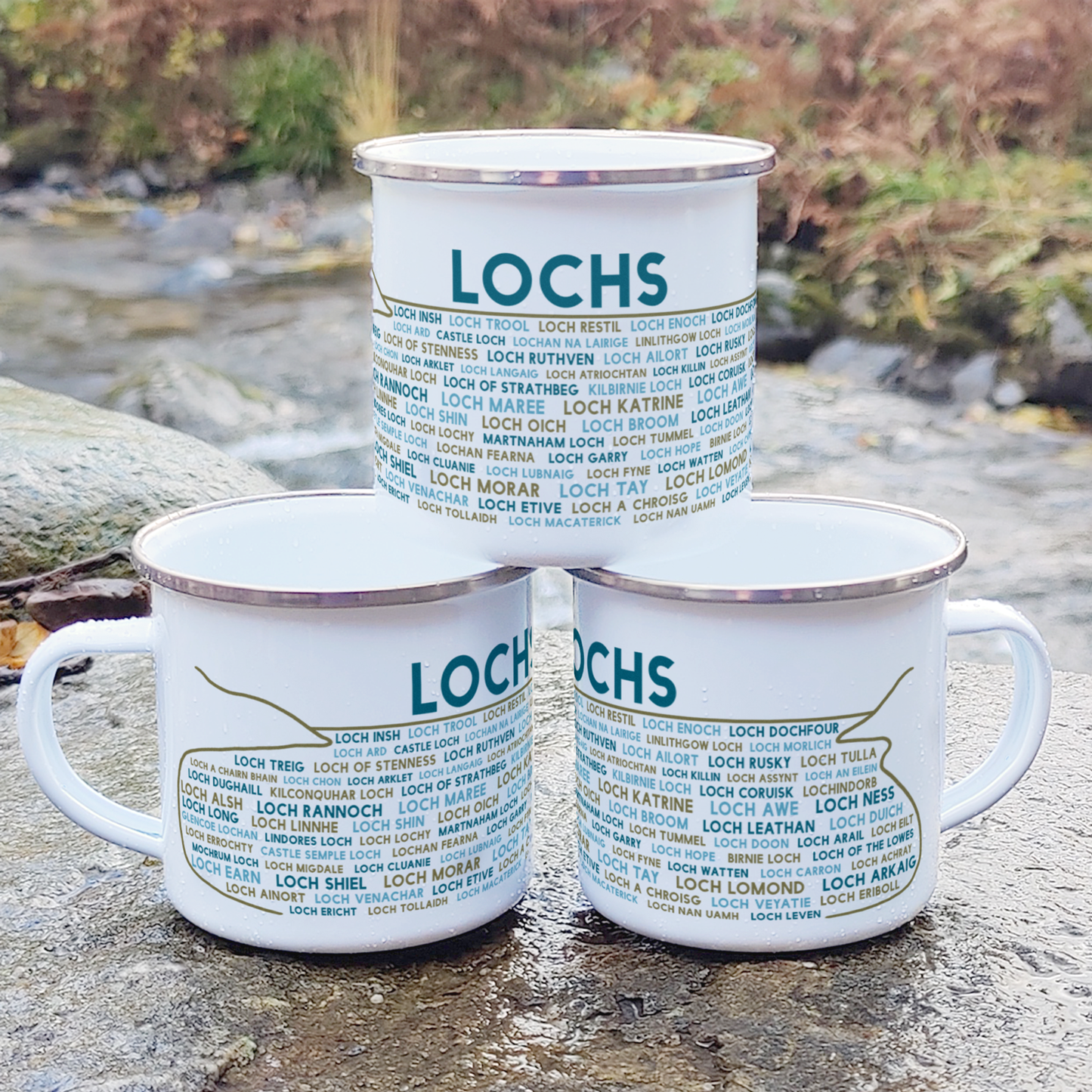 Three stacked white enamel mugs with silver rims, standing on a rock in front of foliage and a river. Each mug features the word "lochs" in blue, with the names of various Scottish lochs underneath it.