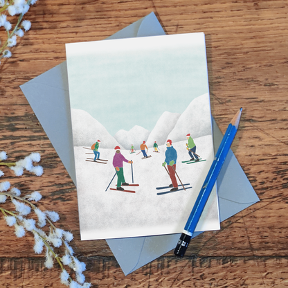 Skiing Christmas card with an illustration of skiers in Santa hats in front of some snowy mountains, laid on a wooden surface with a pencil and some flowers