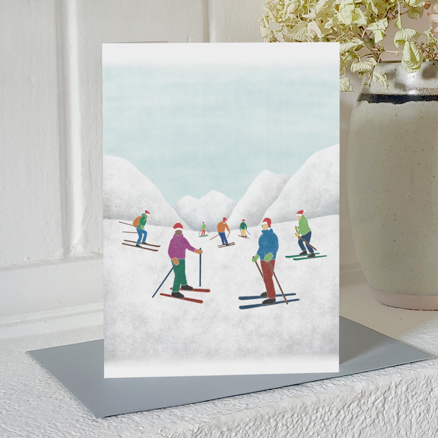 Skiing Christmas card with an illustration of skiers in Santa hats in front of some snowy mountains, standing on a mantelpiece