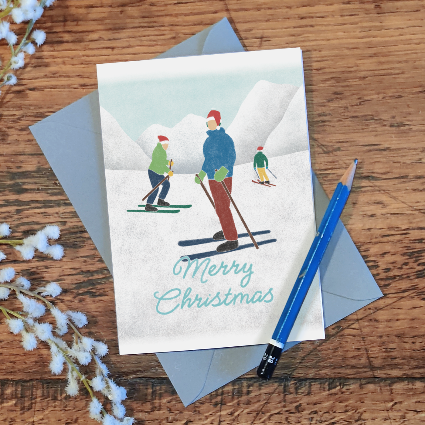 Skiing Christmas card with three skiers in Santa hats and the text 'Merry Christmas', lying on a wooden surface with a pencil and some flowers