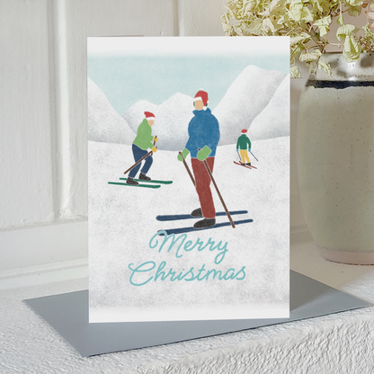 Skiing Christmas card with three skiers in Santa hats and the text 'Merry Christmas', standing on a mantelpiece