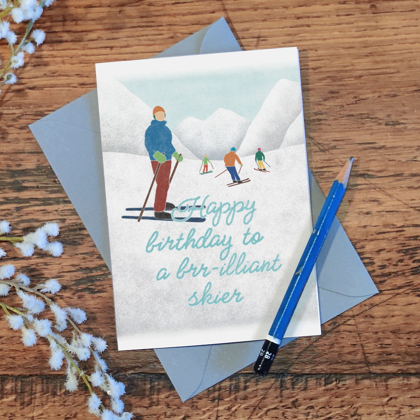 Birthday card with a skiers illustration and the message "happy birthday to a brr-illiant skier", laid on a wooden surface with a pencil and flowers.
