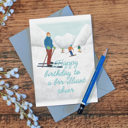 Birthday card with a skiers illustration and the message "happy birthday to a brr-illiant skier", laid on a wooden surface with a pencil and flowers.