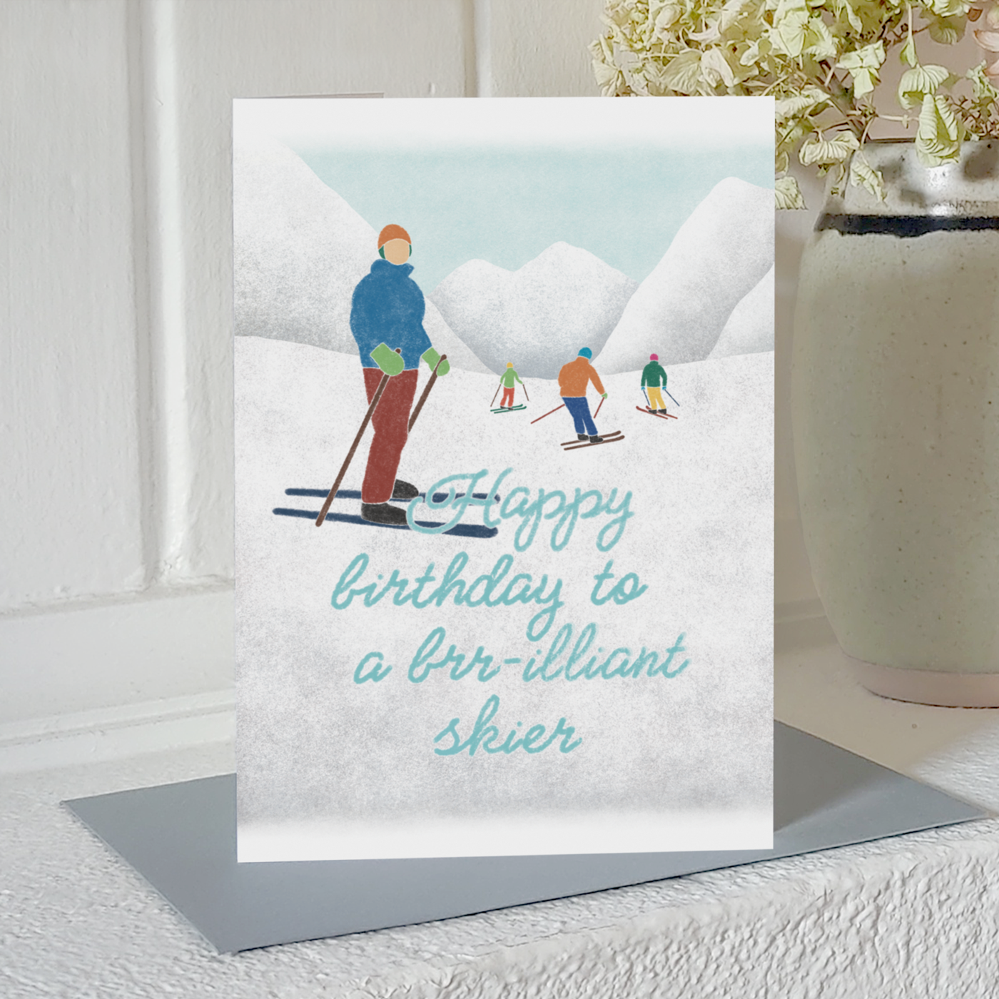 Birthday card with an illustration of four skiers tration and the text "happy birthday to a brr-illiant skier", standing upright on a mantelpiece