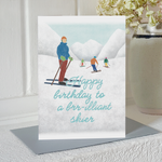 Birthday card with an illustration of four skiers tration and the text "happy birthday to a brr-illiant skier", standing upright on a mantlepiece