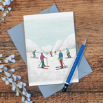 Skiing greetings card, with an illustration of skiers in some snowy mountains, lying on a wooden surface with a pencil and some flowers