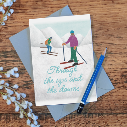 Greetings card with an illustration of skiers and the text 'Through the ups and the downs', lying on a wooden surface with a pencil and flowers