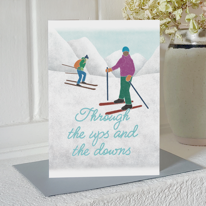 Greetings card with an illustration of two skiers and the text 'Through the ups and the downs' standing up on a mantelpiece