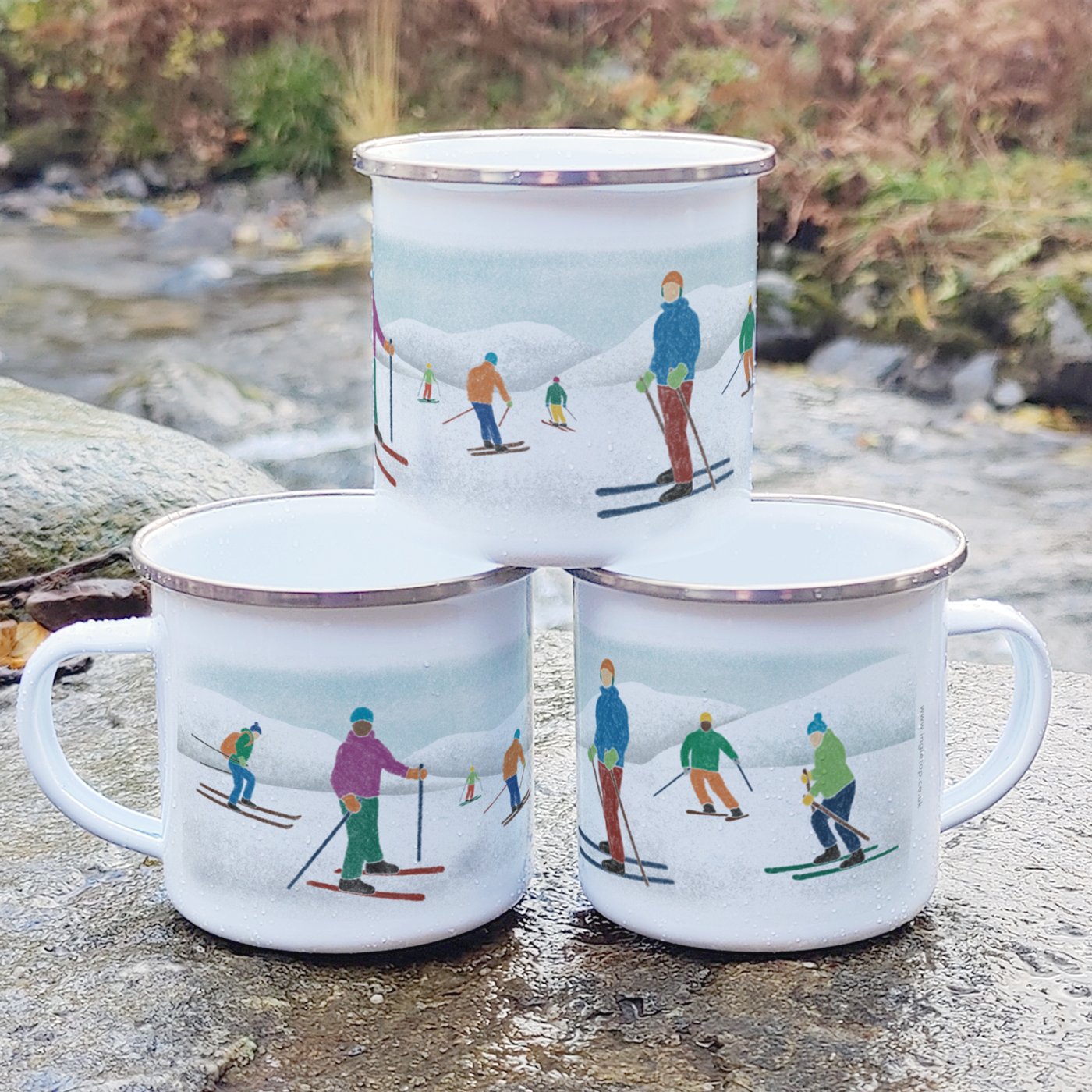 A stack of three enamel mugs printed with our skiing design, standing outdoors on a rock