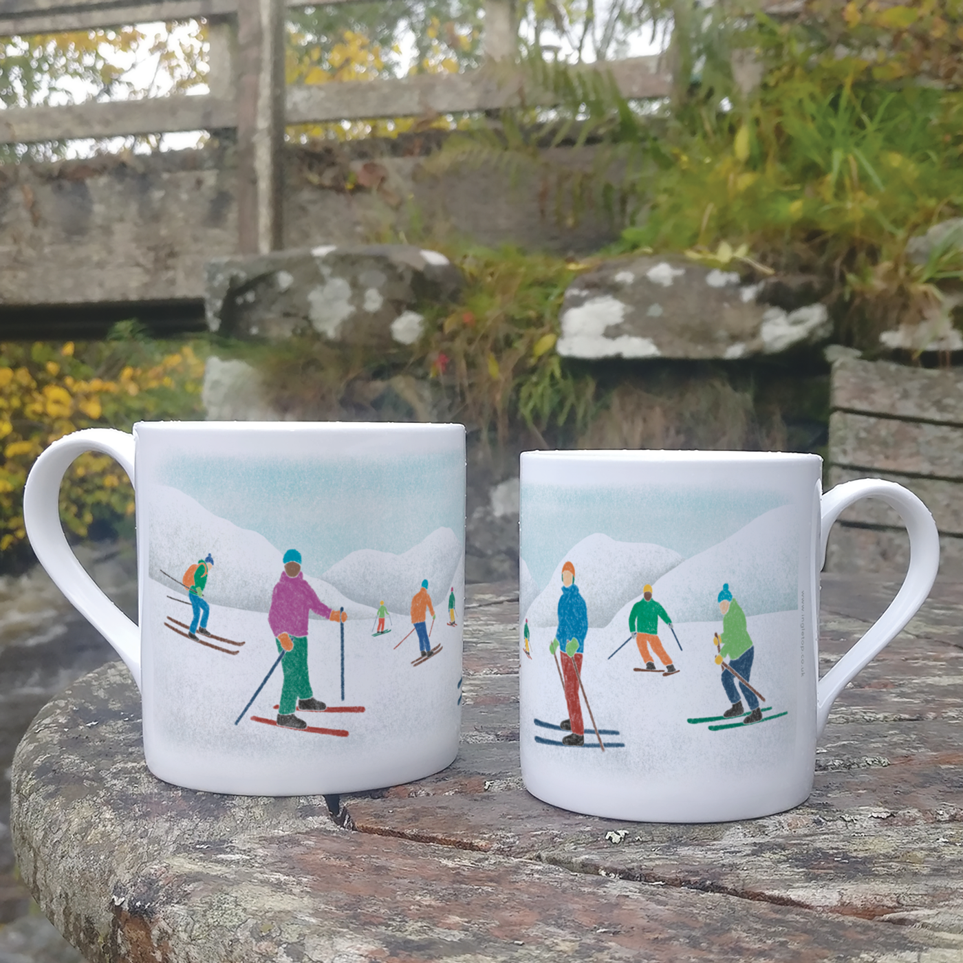Two fine bone china mugs, decorated with with skiing illustrations, on a wooden surface outdoors.