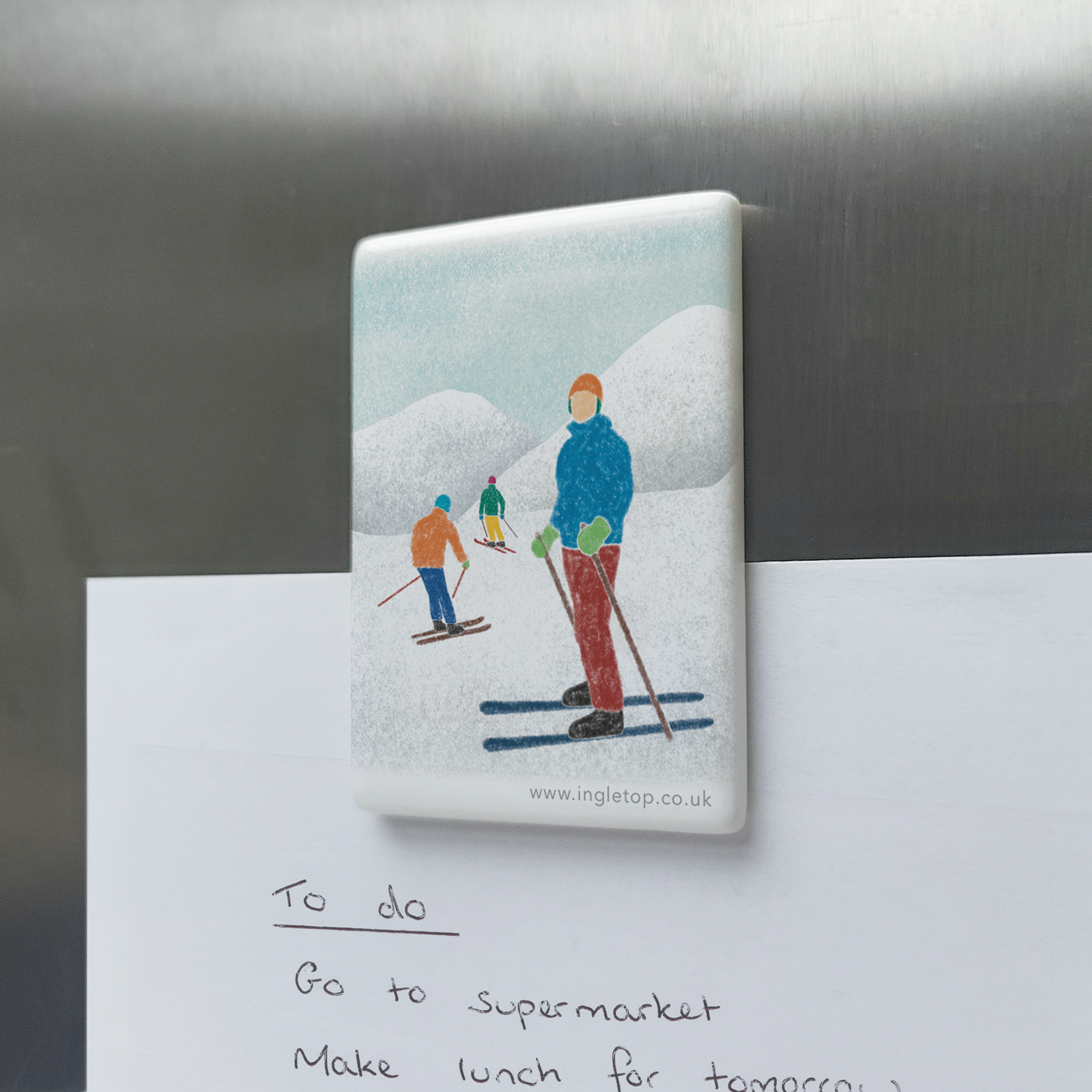 Rectangular ceramic magnet, decorated with an illustration of three skiers in a snowy mountain landscape, holding a paper "to do" list onto a metal surface