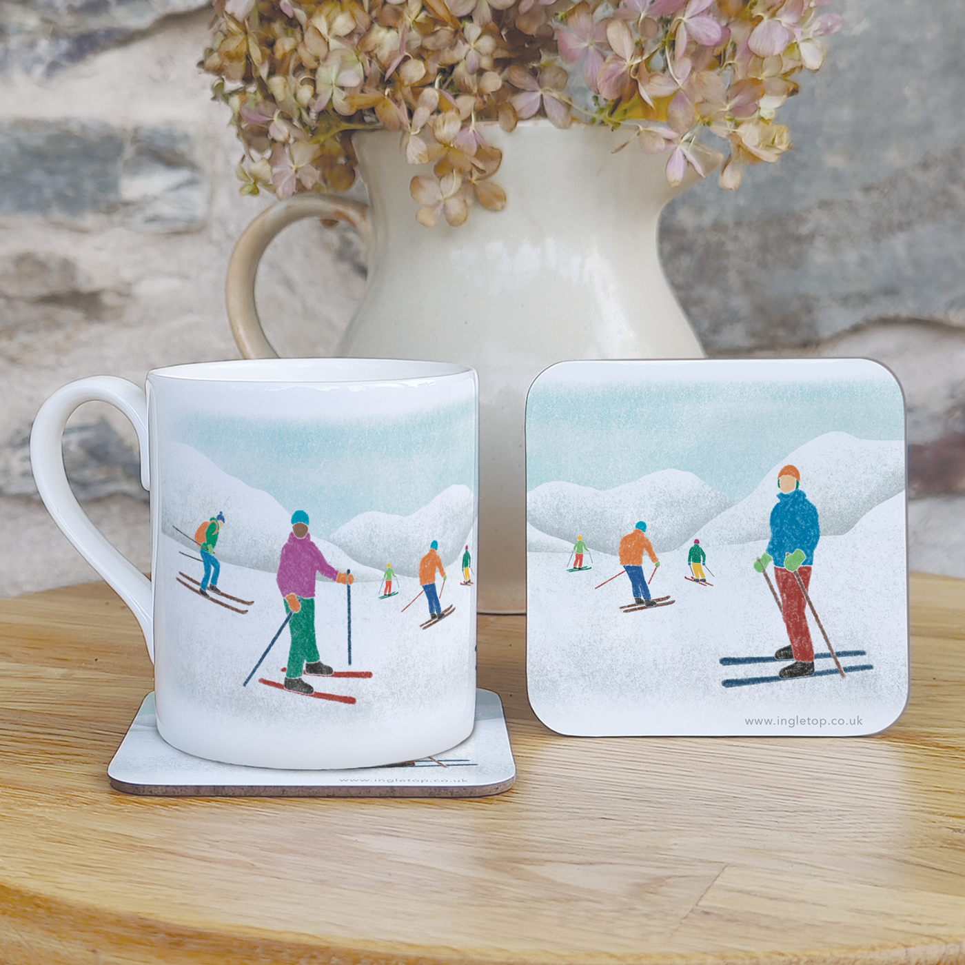 A fine bone china mug and two coasters, decorated with illustrations of skiers in front of snowy mountains, on a wooden table with flowers in the background