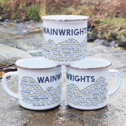 Three stacked white enamel mugs with silver rims, standing on a rock in front of foliage and a river. Each mug features the word "Wainwrights" in purple, with the names of various Lake District fells underneath it.