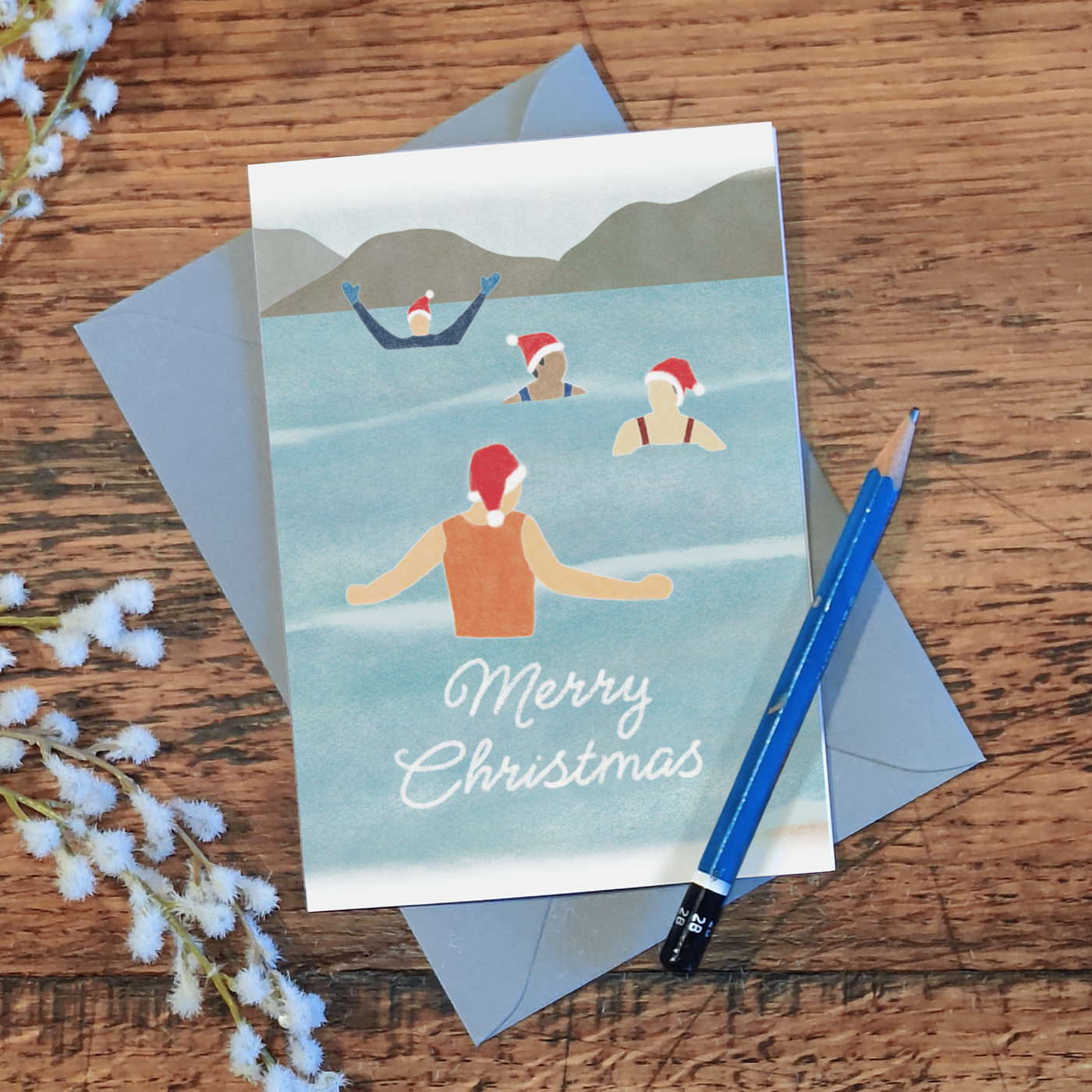 Wild swimming Christmas card with four festive swimmers and the text "Merry Christmas", lying on a wooden surface with a pencil and some flowers