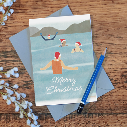 Wild swimming Christmas card with four festive swimmers and the text "Merry Christmas", lying on a wooden surface with a pencil and some flowers