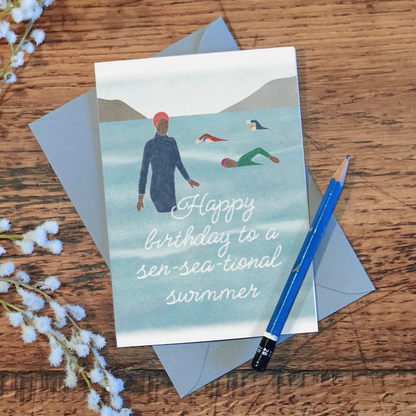 Birthday card with illustration of people ope-water swimming and the text 'Happy birthday to a sen-sea-tional swimmer', laid flat on a wooden surface with a pencil and flowers.