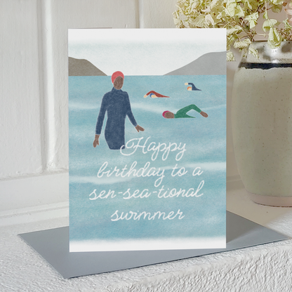 Greetings card with an illustration of people cold water swimming and the text 'Happy birthday to a sen-sea-tional swimmer', standing upright on a mantlepiece